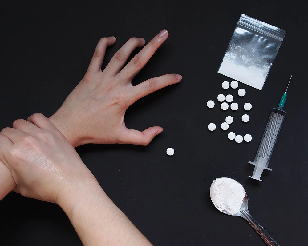 Persoon trekt een hand terug naast ketamine, pillen en een injectiespuit op tafel. De afbeelding sluit aan bij de symptomen van ketaminegebruik en problematisch middelengebruik.