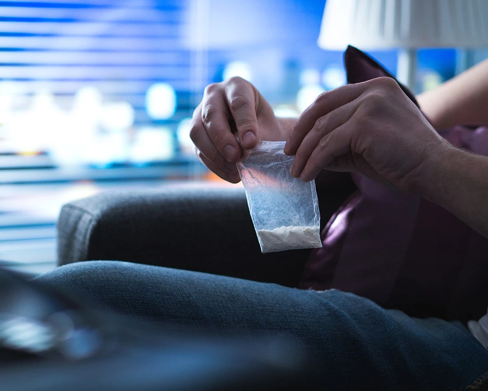 Een persoon houdt een klein plastic zakje met wit poeder vast in een donkere ruimte, passend bij drugsgebruik en verslavingsproblematiek.