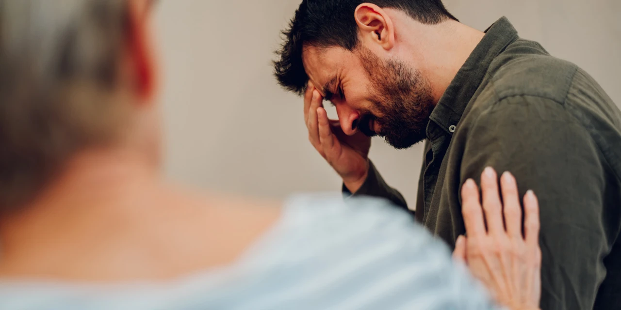 Een man houdt zijn hoofd vast terwijl iemand hem steun geeft tijdens een emotioneel gesprek over alcoholproblemen.