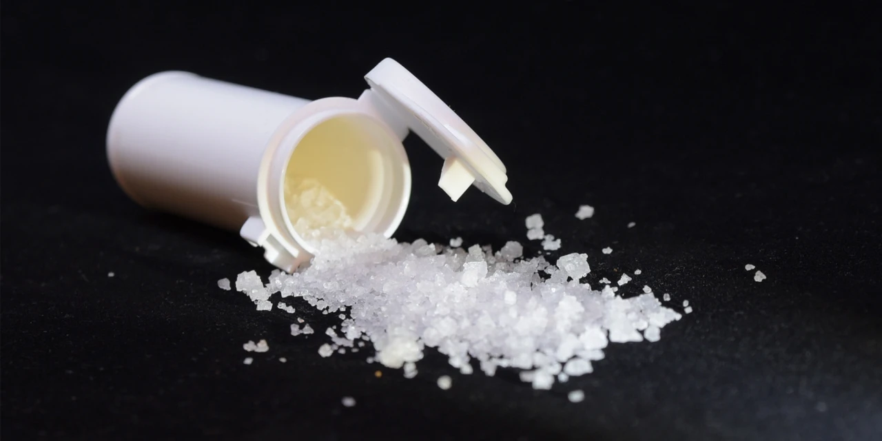 Witte crystal meth kristallen liggen naast een geopend medicijnpotje op een donkere ondergrond. De afbeelding toont crystal meth in kristalvorm en verwijst naar de risico’s van gebruik en verslaving.