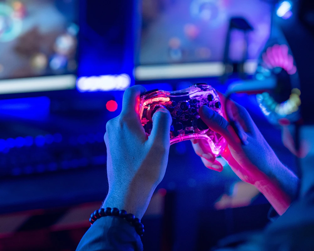 Iemand houdt een verlichte gamecontroller vast terwijl er op schermen videogames zichtbaar zijn. De afbeelding laat gamen zien en verwijst naar mogelijke kenmerken van een gameverslaving.