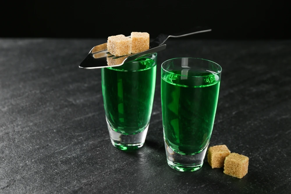 Twee glazen groene absint met suikerklontjes en een absintlepel op tafel.
