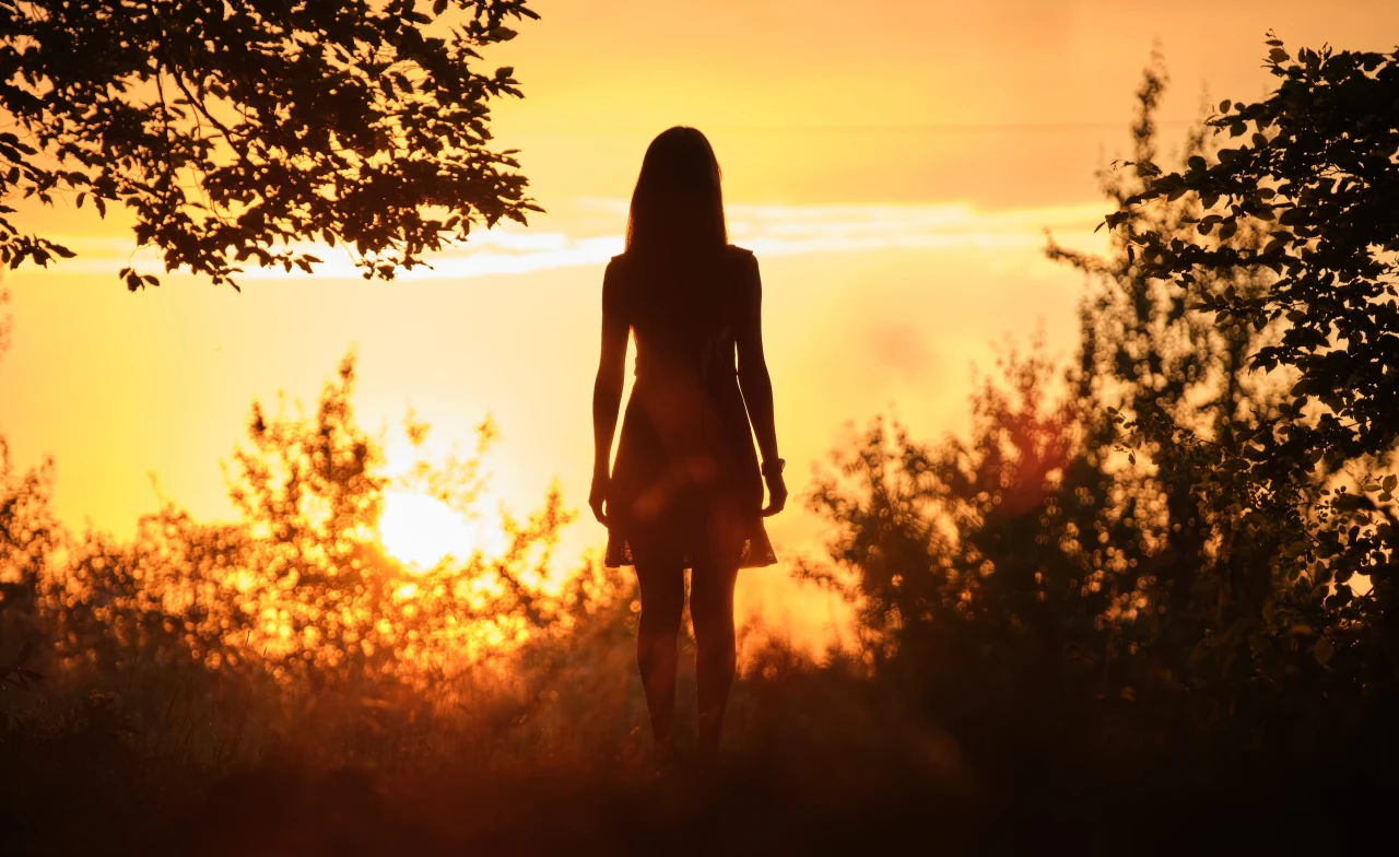 Vrouw staat in bos richting zon te kijken, met haar rug naar de camera.