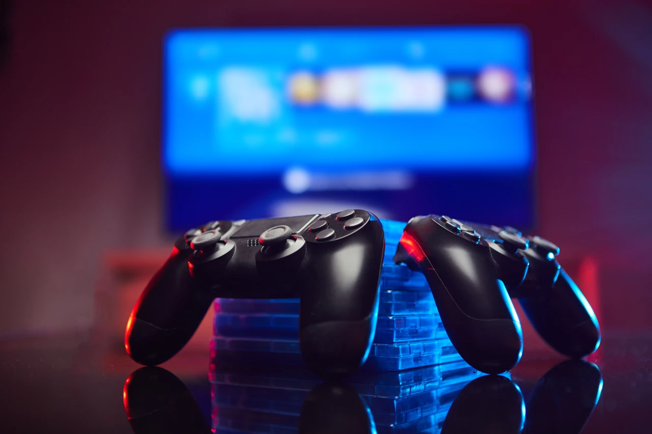 2 zwarte game-controllers liggen op tafel voor een beeldscherm.