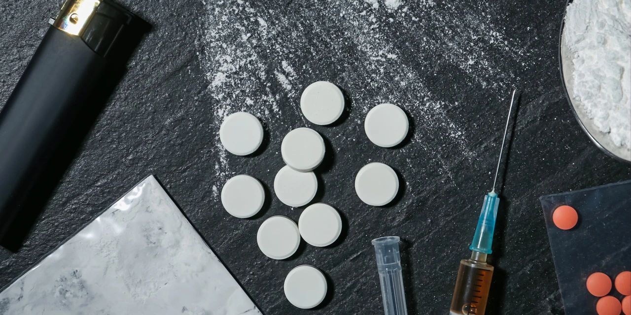 XTC pillen, cocaïnepoeder en een injectiespuit op een donkere ondergrond, passend bij gecombineerd drugsgebruik.