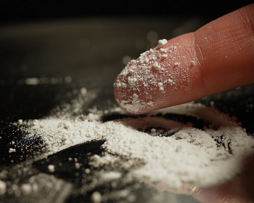 Wit poeder van designerdrugs ligt op een vingertop en verspreid over een donkere ondergrond. De afbeelding laat het gebruik van designerdrugs zien en verwijst naar de risico’s van onbekende samenstellingen.