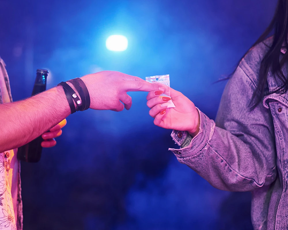 Persoon geeft een zakje met drugs door tijdens een feest of uitgaanssituatie. De afbeelding past bij informatie over XTC effecten en de risico’s van XTC-gebruik.