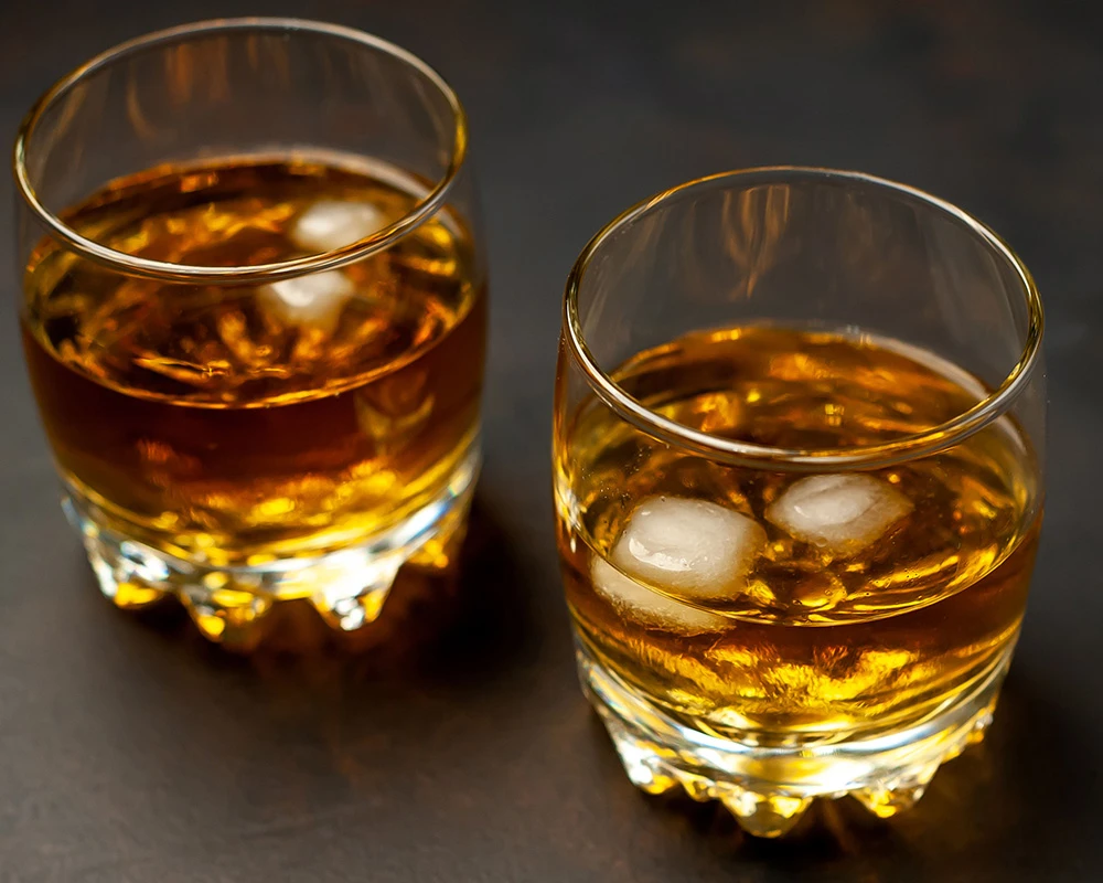 Twee glazen whisky met ijsblokjes staan op een donkere tafel, passend bij alcoholgebruik en alcoholverslaving.