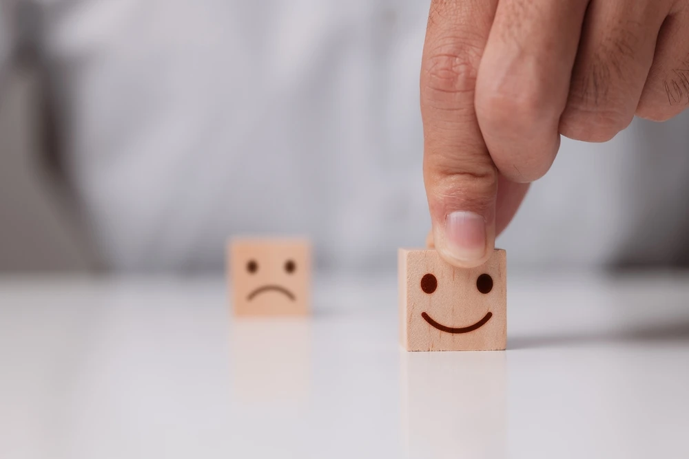 blokje-met-droevig-gezichtje-en-blokje-met-lachend-gezichtje Hand plaatst houten blokje met lachend gezicht voor een houten blokje met een droevig gezicht.