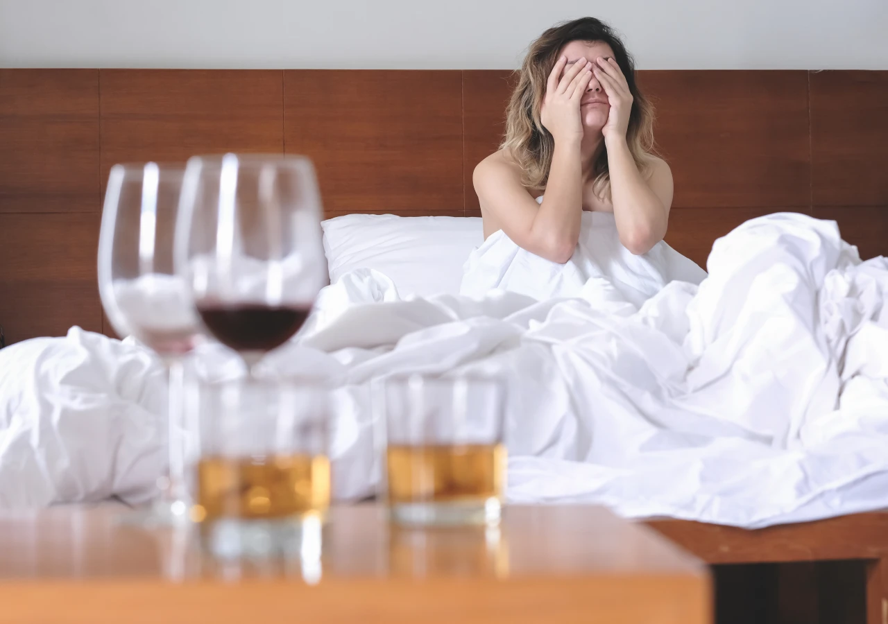 vrouw-zit-in-bed-en-houdt-handen-voor-gezicht-met-op-achtergrond-halflege-glazen-drank Vrouw in bed met handen voor gezicht en halflege glazen op tafel en voelt de gevolgen van alcohol.