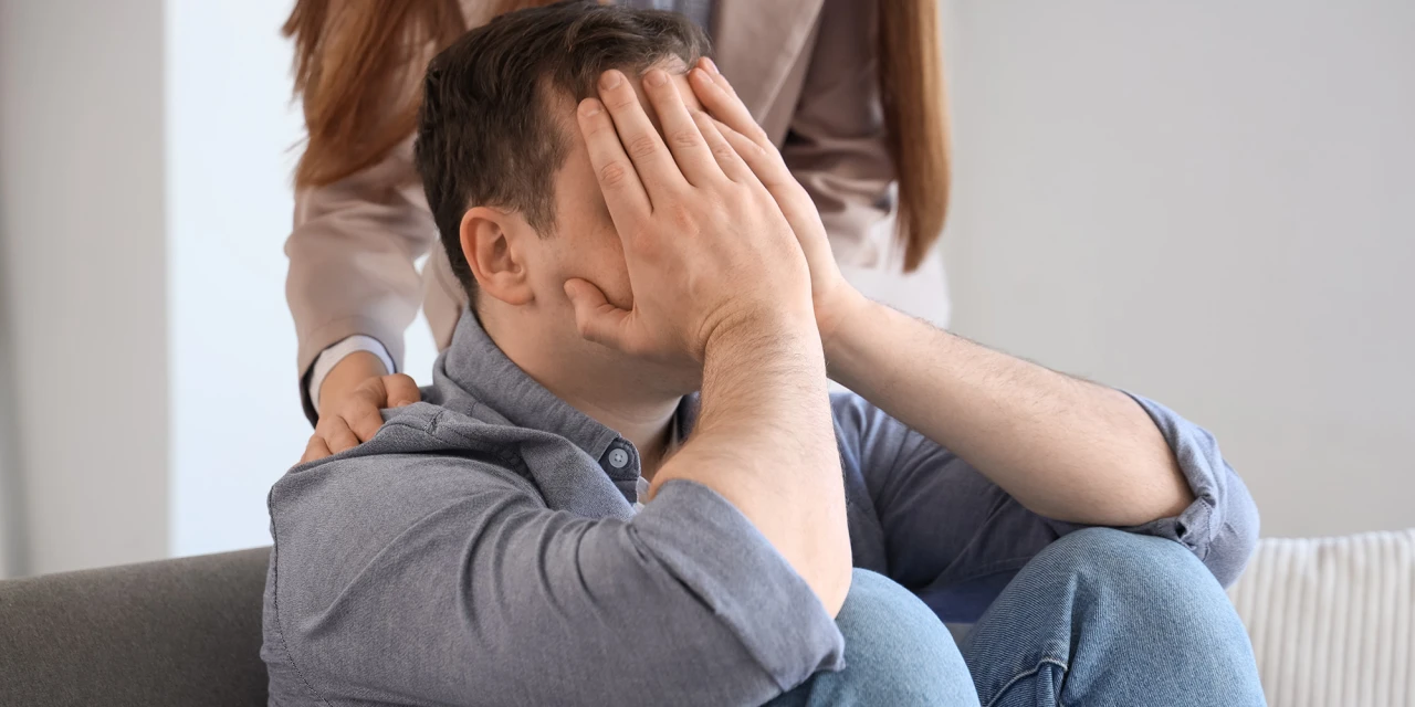 Een man zit verdrietig met zijn handen voor zijn gezicht terwijl iemand steun biedt.
