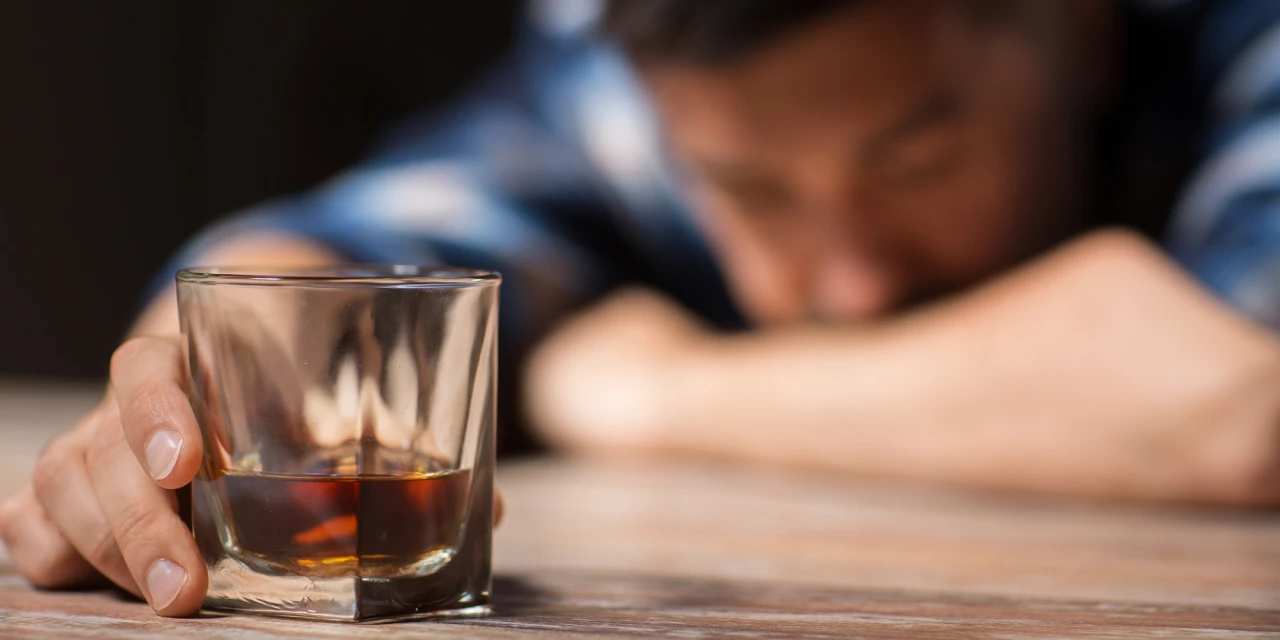 Een persoon ligt vermoeid aan tafel met een glas sterke drank na een periode van overmatig alcoholgebruik.
