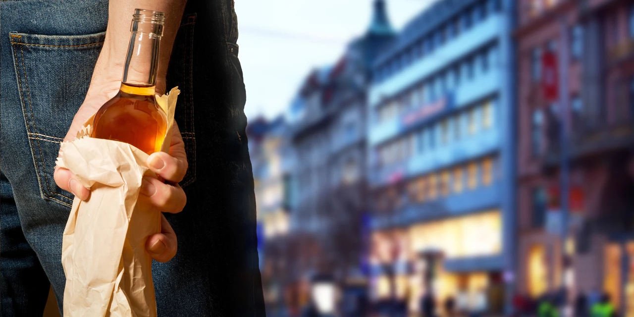 Een persoon houdt een fles alcohol in een papieren zak vast terwijl hij op straat loopt, wat kan wijzen op verborgen alcoholgebruik.