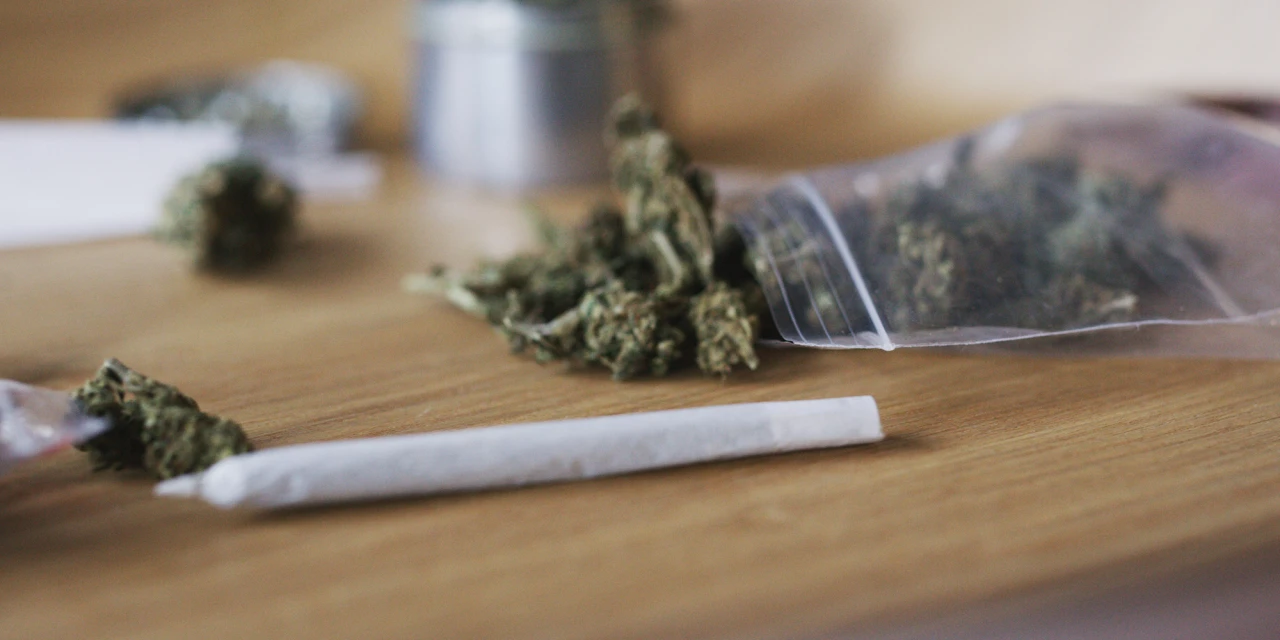 Een joint ligt op een houten tafel naast cannabis en een open zakje met wiet.