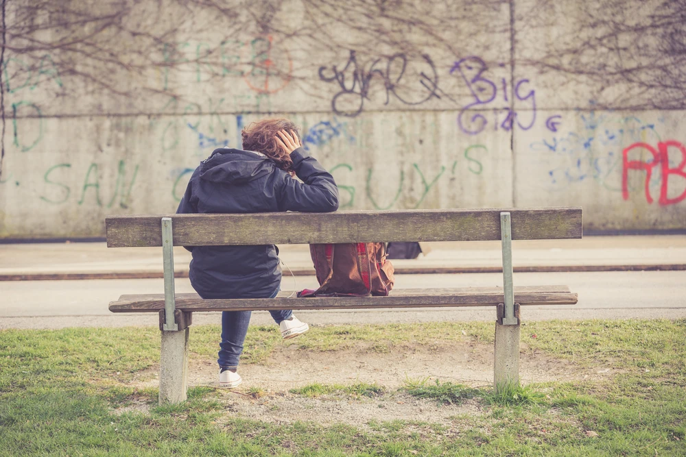 Iemand zit op een bankje tegen een grijze muur met graffiti aan te kijken en is depressief doordat blowen zijn leven kapot maakt. 