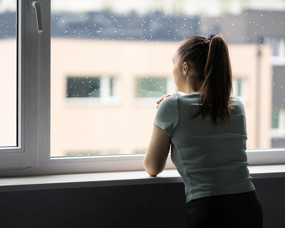Een vrouw kijkt naar buiten terwijl ze alleen voor een raam staat. De afbeelding verwijst naar gevoelens van isolatie, verwarring en mentale klachten die kunnen ontstaan bij een psychose door drugs.