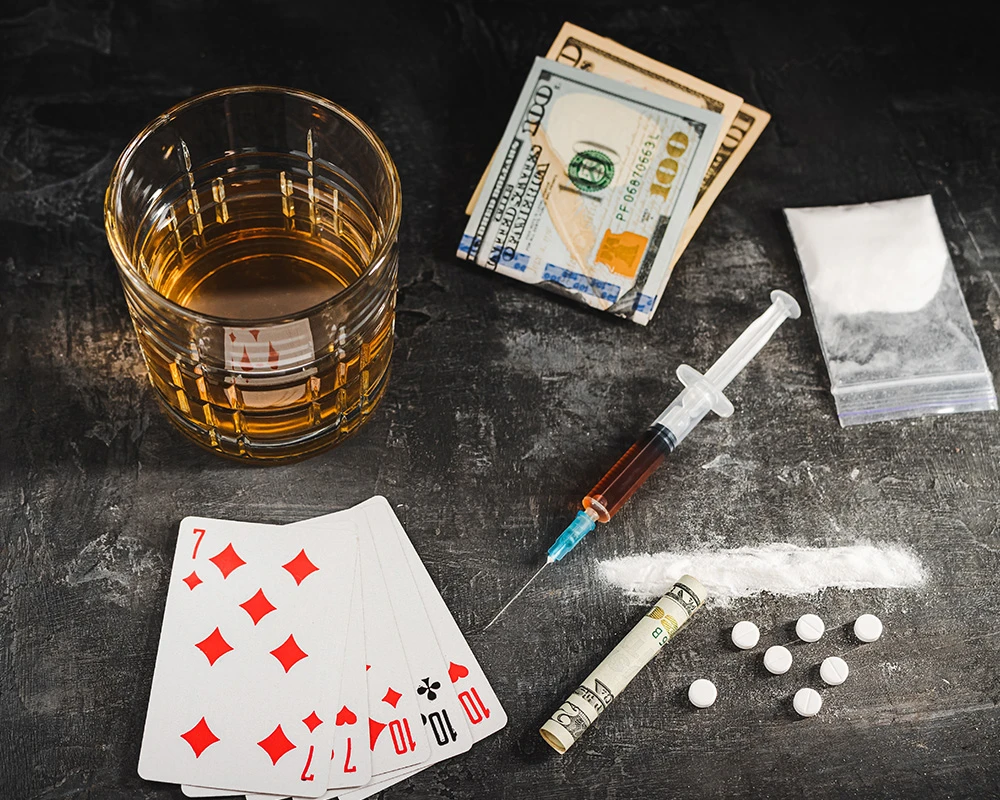 Speelkaarten, alcohol, geld en cocaïne liggen samen op tafel naast gokattributen. De afbeelding verwijst naar de combinatie van gokken, drugsgebruik en risicovol gedrag.