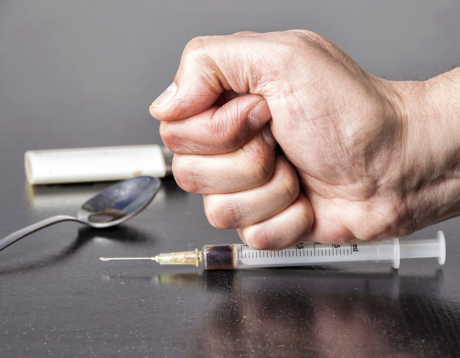 Gebalde vuist naast een injectiespuit en drugsattributen als symbool voor het willen stoppen met middelengebruik en het overwinnen van een verslaving.
