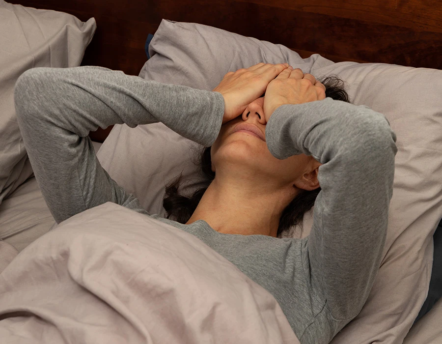 Een vrouw ligt wakker in bed met haar handen voor haar gezicht. De afbeelding laat vermoeidheid en slaapproblemen zien die kunnen ontstaan door een plafonddienst.