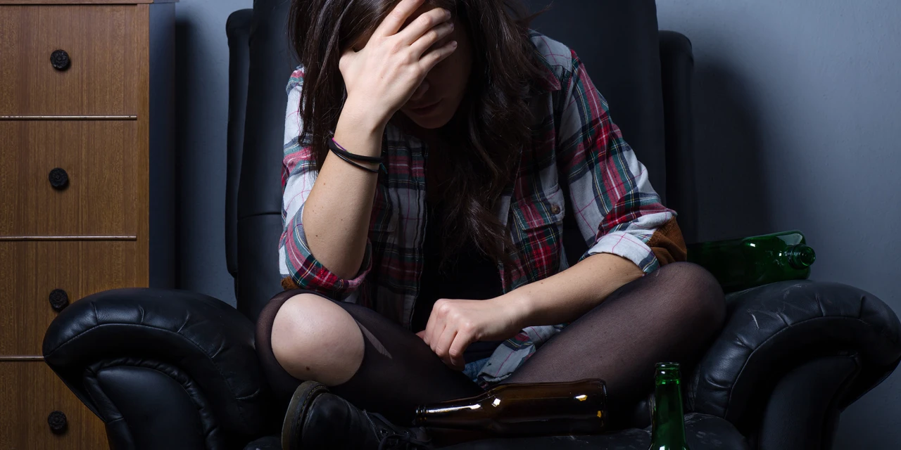 Een vrouw zit somber in een stoel tussen lege alcoholflessen, wat kan wijzen op de mentale en fysieke gevolgen van langdurig alcoholgebruik.