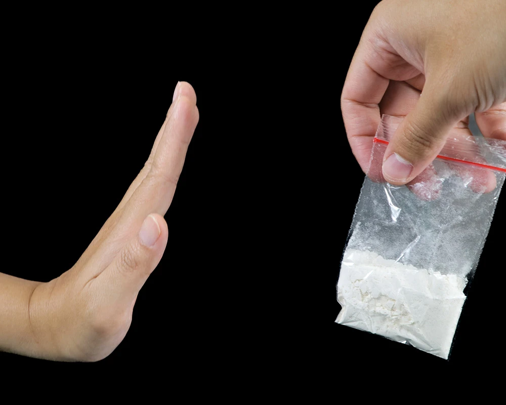 Een hand houdt een zakje met wit poeder vast terwijl een andere hand een afwerend gebaar maakt. De afbeelding symboliseert het weigeren van drugs en het maken van een bewuste keuze.