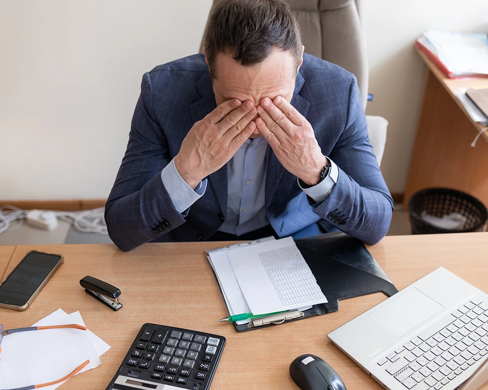 Man in pak zit gestrest achter een bureau met documenten, laptop en rekenmachine. De afbeelding laat de emotionele en financiële gevolgen van gokverslaving zien.