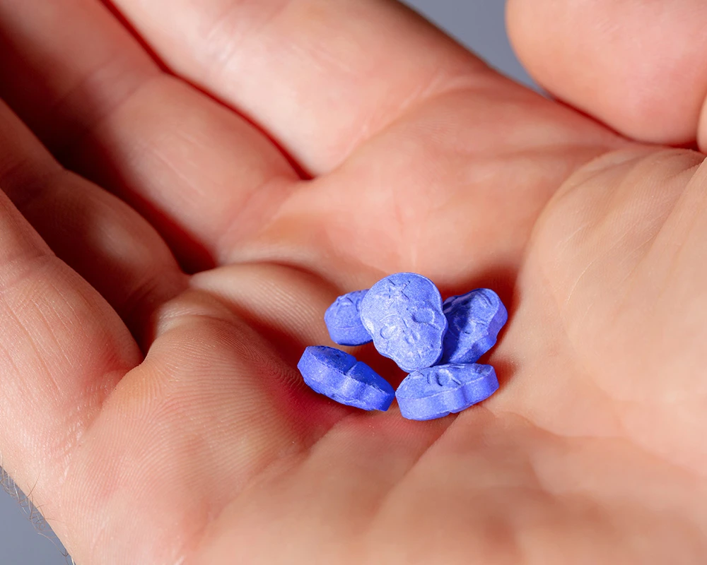 Hand met meerdere blauwe MDMA pillen close-up in beeld. De afbeelding past bij recreatief drugsgebruik en de risico’s van een mdma verslaving.