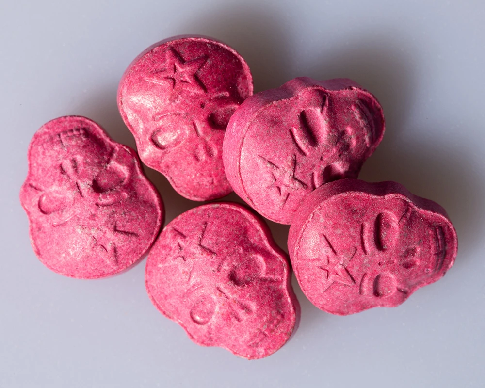 Meerdere roze XTC-pillen in schedelvorm liggen bij elkaar op een lichte achtergrond. De afbeelding hoort bij informatie over wat is XTC en de risico’s van XTC-gebruik.