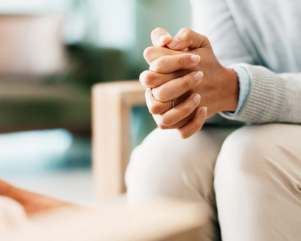 Persoon zit met gevouwen handen tijdens een gesprek over herstel en begeleiding bij verslaving. De afbeelding hoort bij hulp en ondersteuning binnen de verslavingszorg.