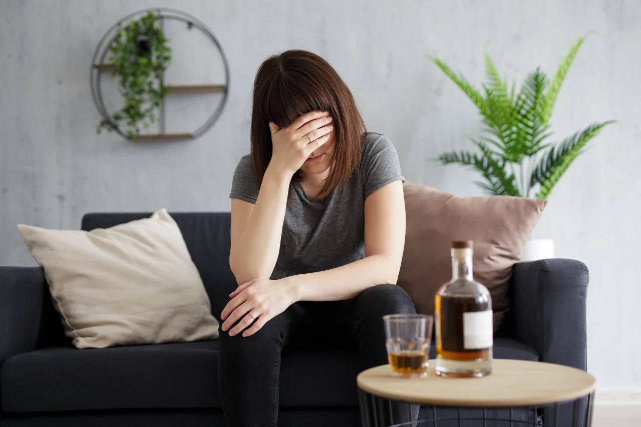 vrouw-zit-met-hand-voor-gezicht-en-fles-drank-op-tafel-in-bank Vrouw zit met fles drank en halfleeg glas met hand voor gezicht in de bank.