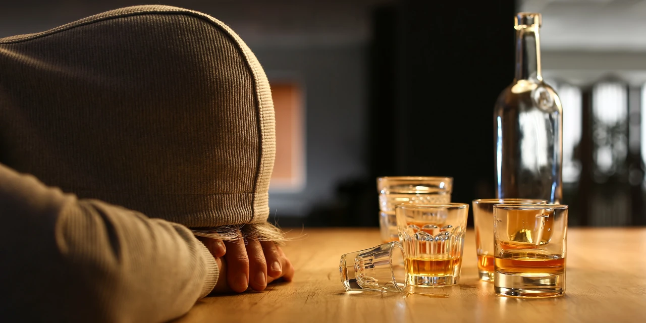 Een persoon ligt met het hoofd op tafel naast meerdere glazen en flessen alcohol tijdens een moment van overmatig alcoholgebruik.