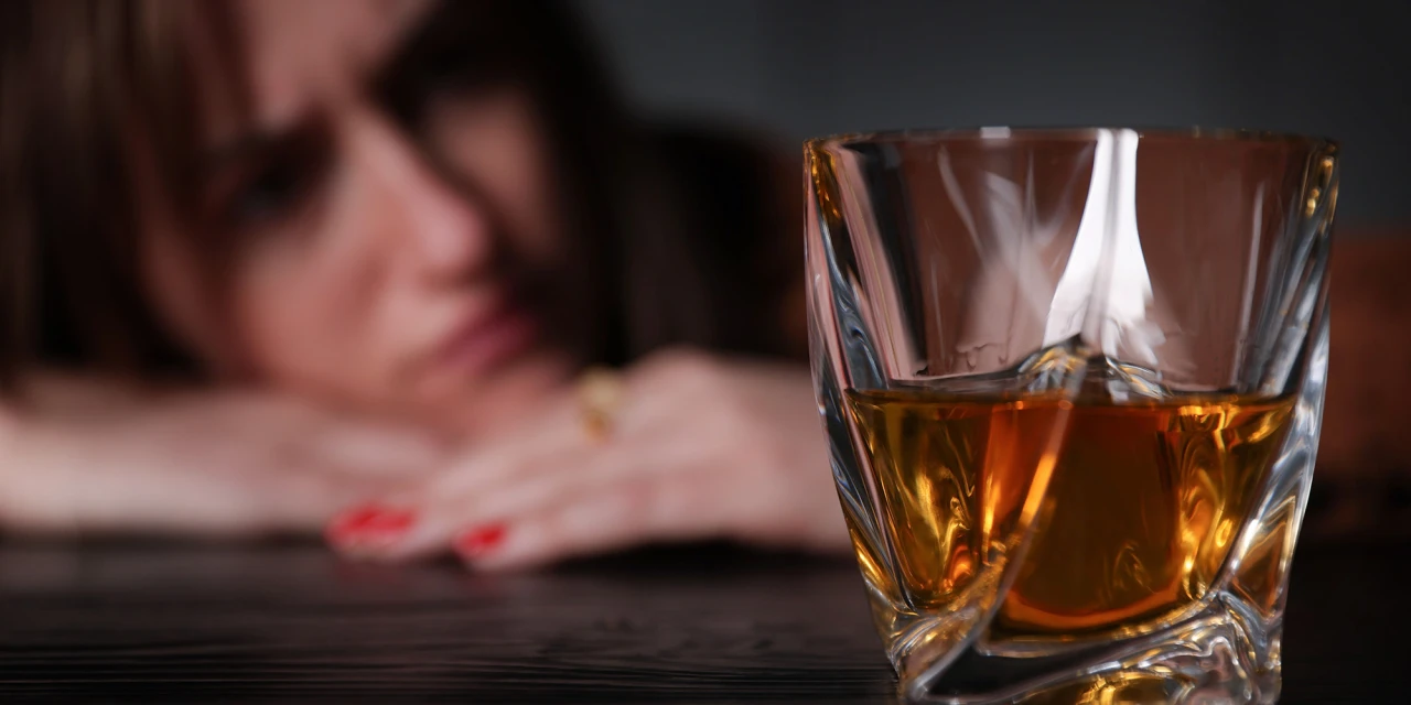Een vrouw kijkt vermoeid en somber naar een glas alcohol dat op tafel staat.