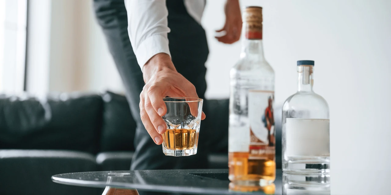 Een persoon pakt een glas sterke drank van tafel terwijl meerdere flessen alcohol zichtbaar zijn.