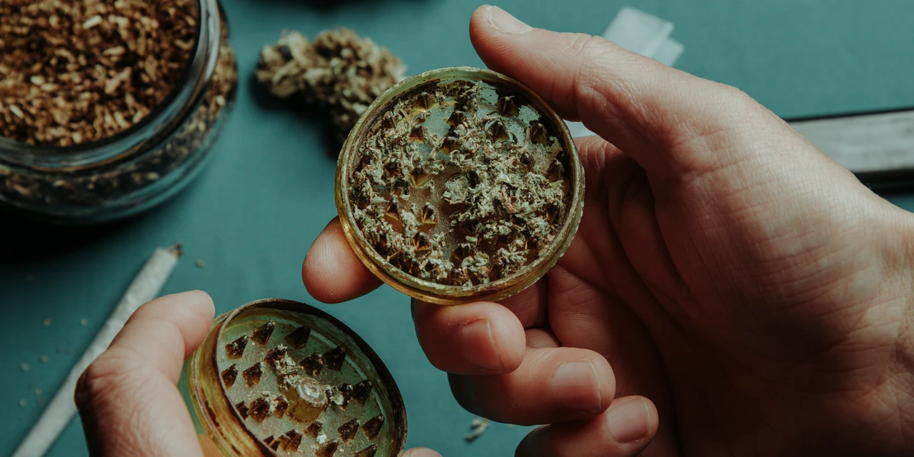 Iemand houdt een grinder met gemalen cannabis vast terwijl er wiet en een joint op tafel liggen.