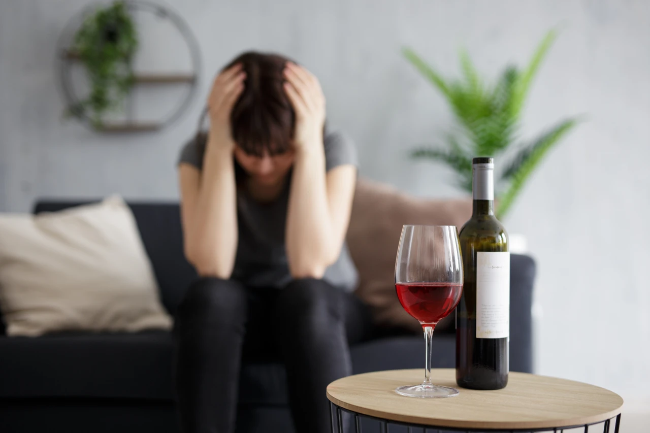 wanhopige-vrouw-bij-fles-wijn Vrouw zit op de bank met hoofd in handen na terugval door alcohol; open fles wijn staat voor haar.