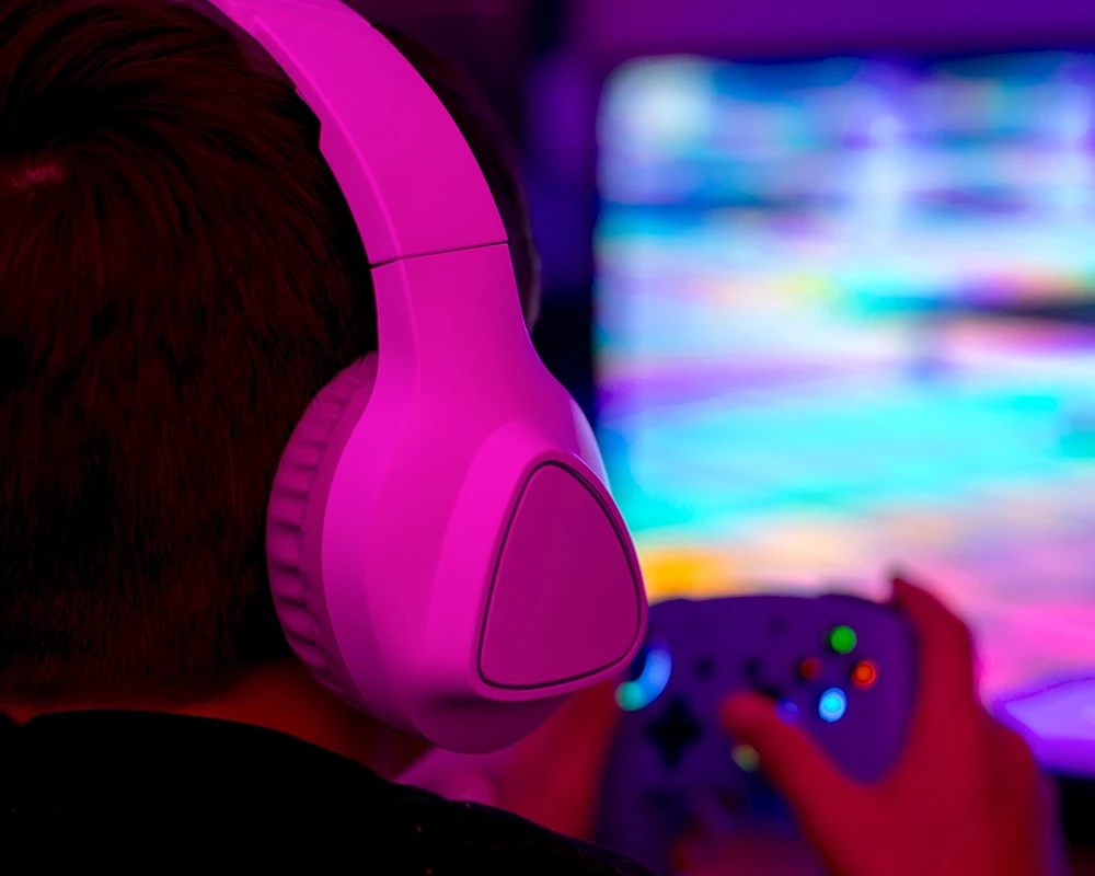 Iemand draagt een headset en speelt een videogame met controller voor een verlicht scherm. De afbeelding verwijst naar langdurig gamen en mogelijke kenmerken van verslaafd zijn aan League of Legends.