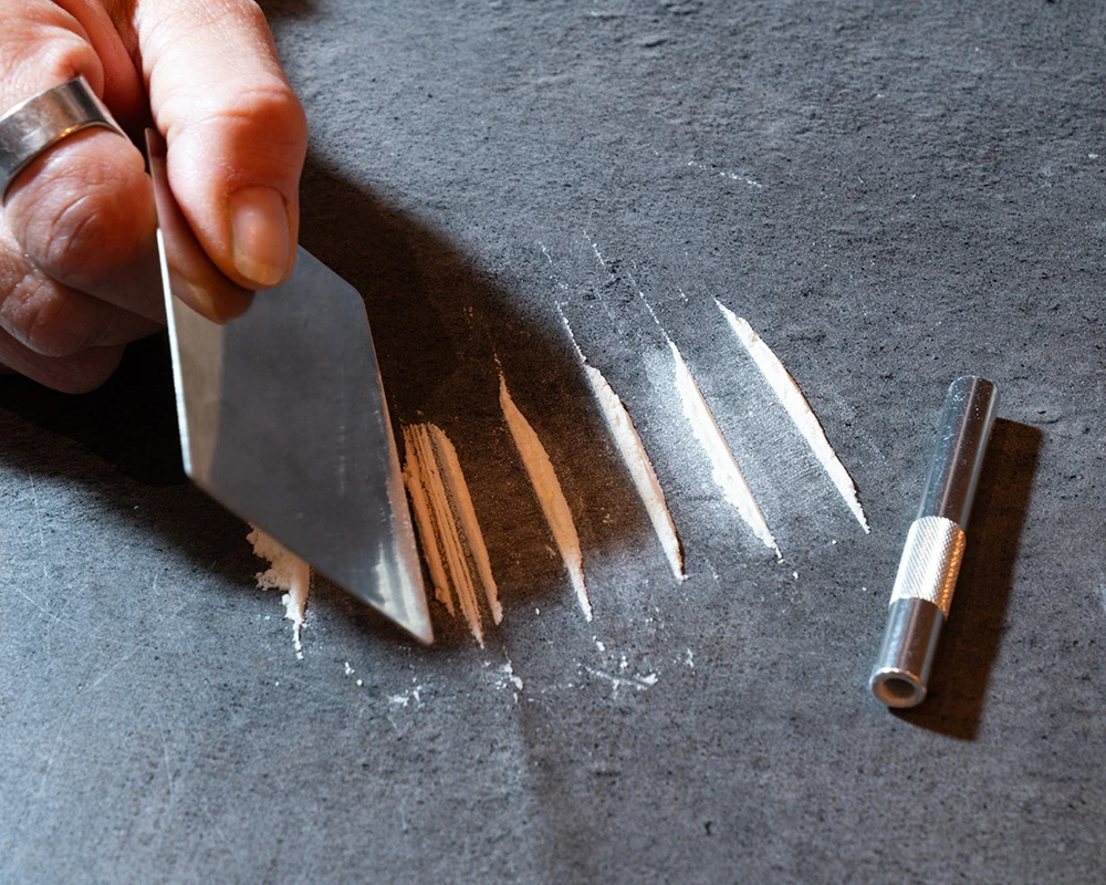 Hand maakt lijnen van wit poeder klaar naast een buisje op tafel. De afbeelding verwijst naar ketaminegebruik en de risico’s op lichamelijke klachten zoals ketamine blaasproblemen.