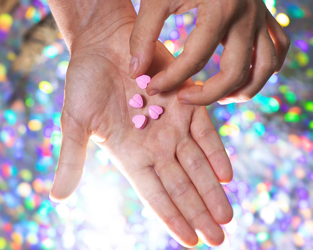 Hand met meerdere roze MDMA pillen in beeld tegen een kleurrijke achtergrond. De afbeelding laat recreatief drugsgebruik zien en past bij informatie over wat is mdma.