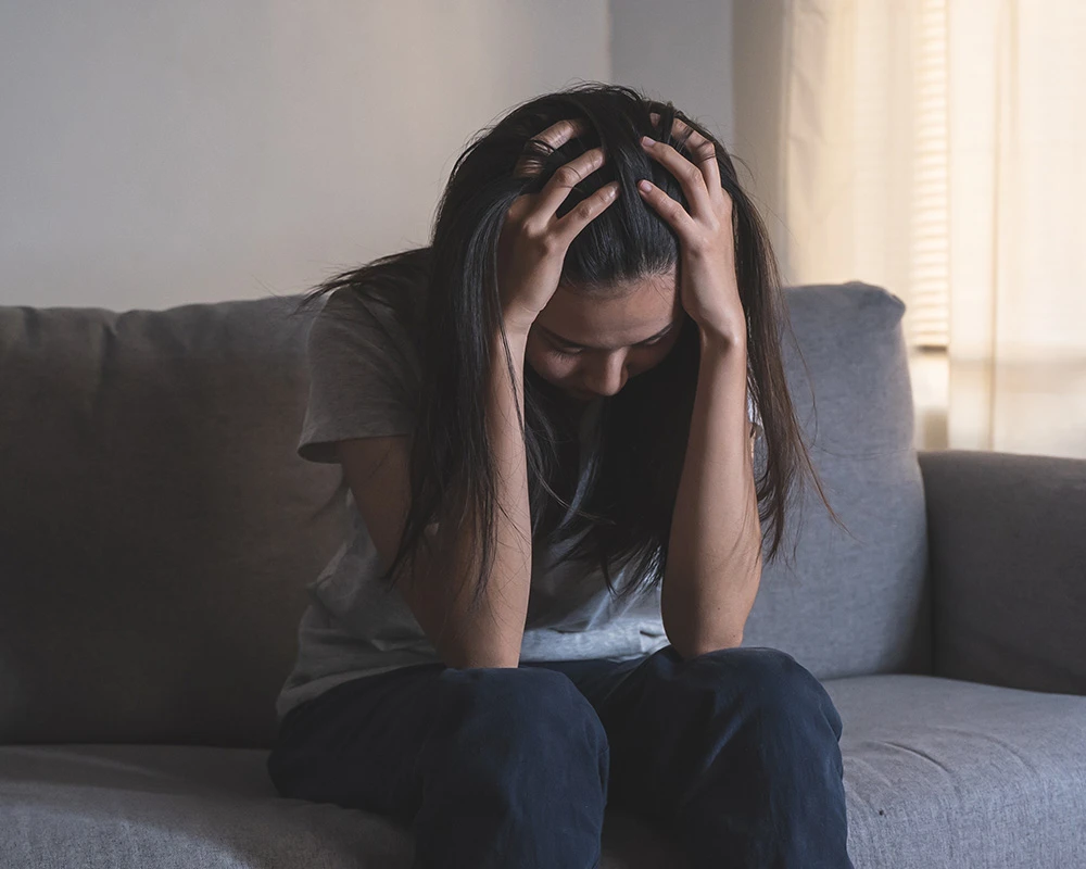 Vrouw zit somber op de bank met haar handen in het haar. De afbeelding past bij informatie over afkickverschijnselen XTC en mentale klachten na gebruik.