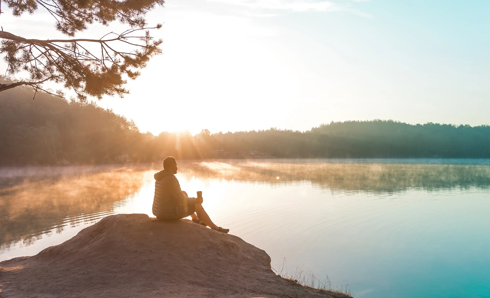 man-drinkt-koffie-aan water-bij-zonnig-weer Man zit op heuveltje aan het water en drinkt koffie terwijl de zon schijnt.