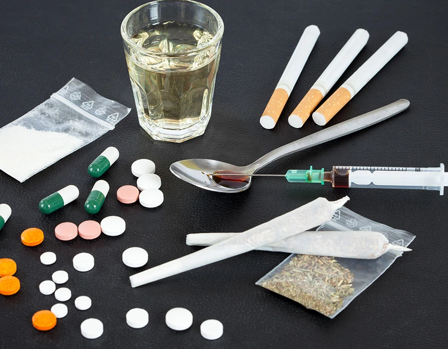 Verschillende soorten middelen zoals pillen, alcohol, joints, poeder en een spuit liggen verspreid op tafel. De afbeelding laat het verschil tussen hard en soft drugs en verschillende vormen van drugsgebruik zien.