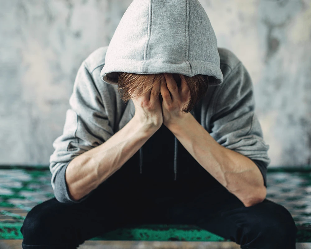 Jonge man met hoodie zit voorovergebogen met zijn handen voor zijn gezicht. De afbeelding laat emotionele klachten en afkickverschijnselen gokken zien.