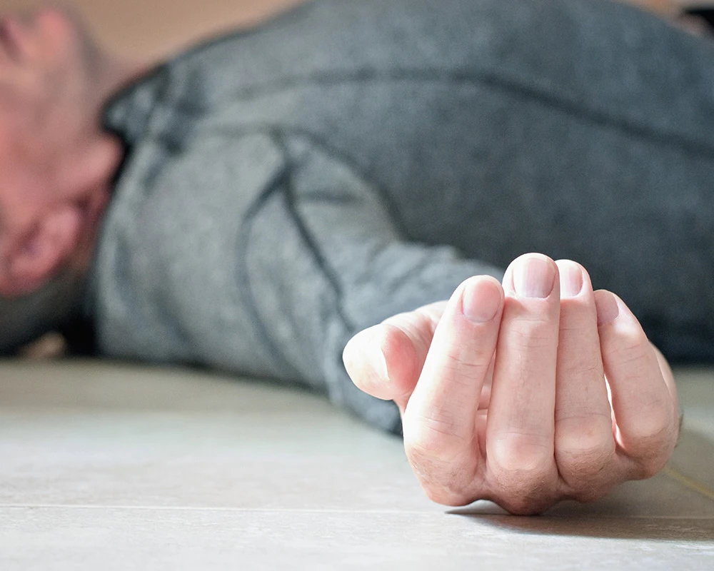 Man ligt bewegingloos op de vloer met een hand op de voorgrond. De afbeelding sluit aan bij een k-hole en de risico’s van overmatig ketaminegebruik.
