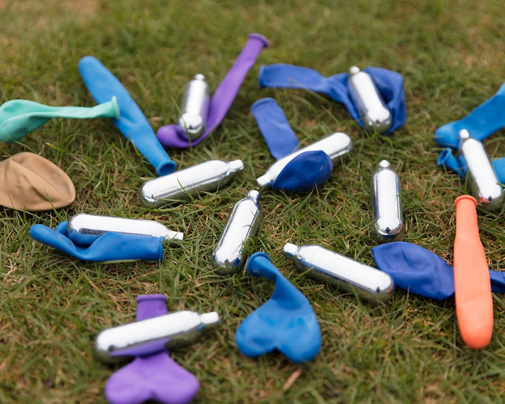 Lachgaspatronen en ballonnen liggen verspreid op een grasveld. De afbeelding verwijst naar recreatief gebruik en de risico’s van een lachgasverslaving.