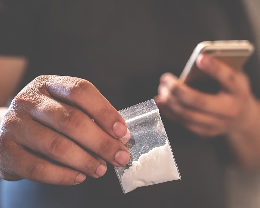 Een persoon houdt een zakje met wit poeder vast terwijl op de achtergrond een telefoon zichtbaar is, passend bij cocaïnegebruik en verslaving.