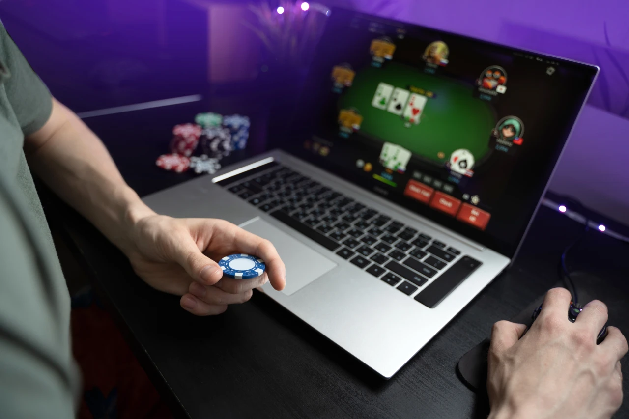 Man houdt fiche vast bij opengeklapte laptop met daarop online kaartspel en legt uit wat gokken is.