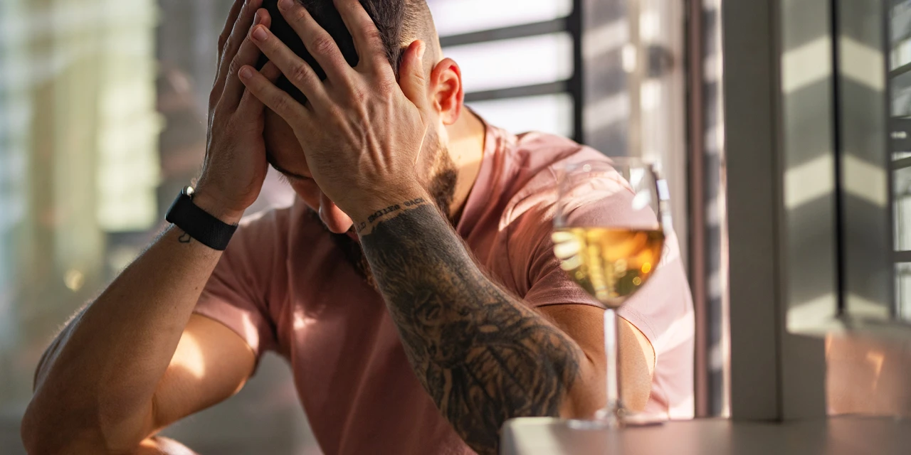Een man zit met zijn handen voor zijn gezicht naast een glas alcohol tijdens een moeilijk moment.