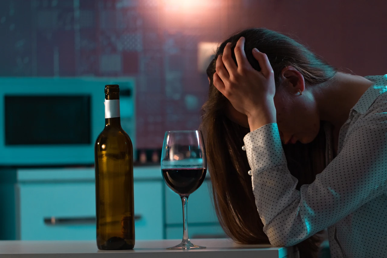 Vrouw aan eettafel maakt zich zichtbaar zorgen, heeft het hoofd in de handen. Op tafel staat een wijnfles en een wijnglas.