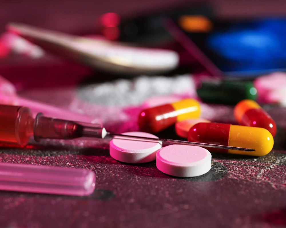 Pillen, capsules, wit poeder en een injectiespuit liggen verspreid op een donkere ondergrond. De afbeelding toont middelengebruik en verwijst naar het verschil tussen hard en soft drugs.