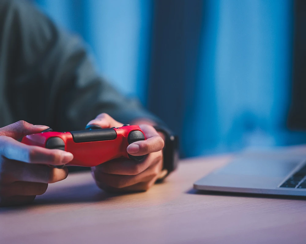 Iemand houdt een rode gamecontroller vast naast een laptop tijdens het gamen. De afbeelding verwijst naar intensief gamen en gedrag dat kan passen bij verslaafd zijn aan Fortnite.